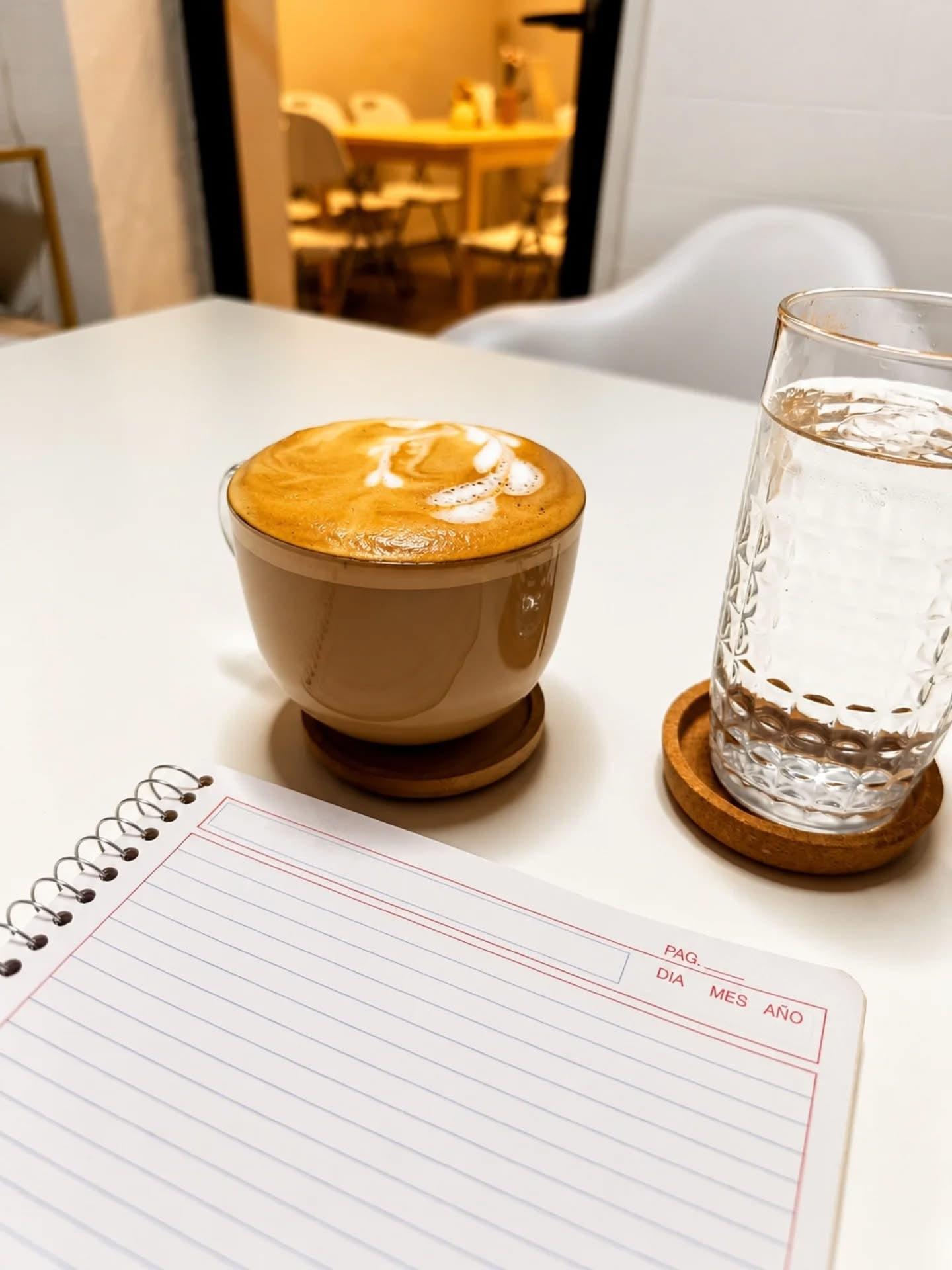 Cappuccino in a ceramic cup next to a glass of water and an open notebook in the Alere Café meeting room